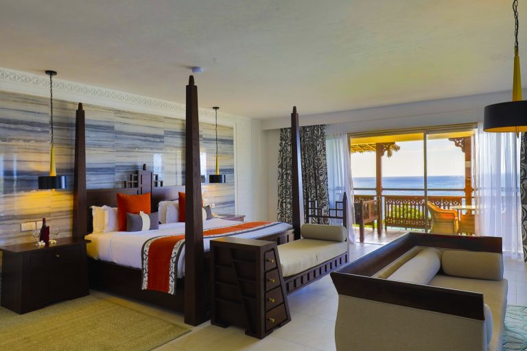 Zimmer der ocean-front-suite mit Himmelbett aus dunkelbraunem Holz, blau-grauen Wandpaneelen und Meerblick vom Balkon