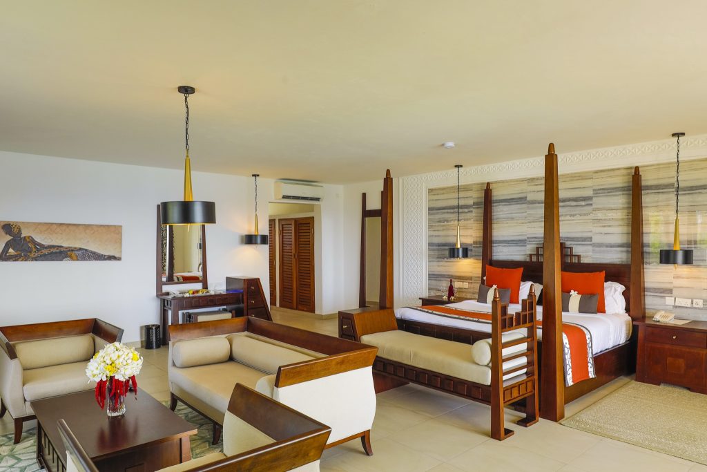 Hotelzimmer mit Himmelbett, dunklen Holzmöbeln, Sofas, Couchtisch und dekorativer Holzwand im Royal Zanzibar Beach Resort