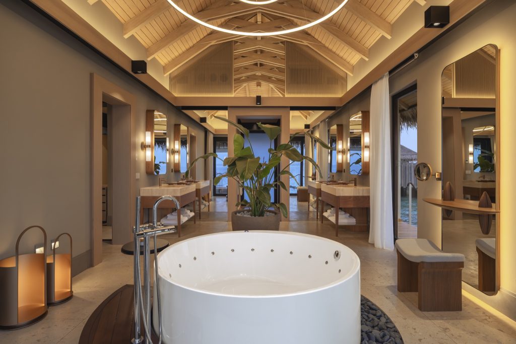 Badezimmer mit runder weißer freistehender Badewanne, Holzgebälk, hellen Steinfliesen und Glasfenstern mit Wasserblick