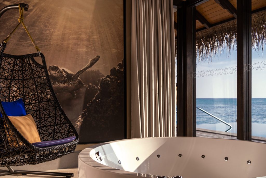 Zimmer der Sunset Deluxe Water Pool Villa mit runder weißer Badewanne, Hängesessel und Meerblick auf den