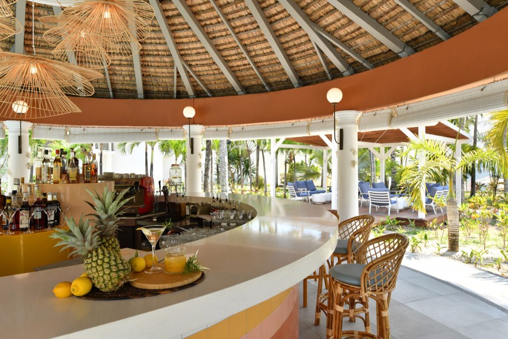 Bar im Veranda Palmar Beach Hotel mit geschwungener Theke, Rattan-Barhockern und Blick auf tropische Pflanzen