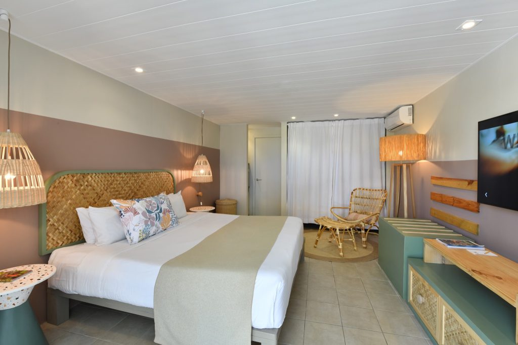 Hotelzimmer mit großem Bett und Kopfteil aus geflochtenem Holz, Kissen mit Blumenmuster, Lampen mit Korblampschirm.
