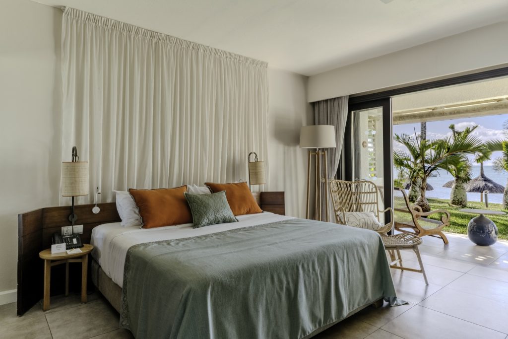 Hotelzimmer Superior Room mit großem Bett, grünem Überwurf, Rattan-Stuhl und Glas-Schiebetüren mit Blick auf Palmen.