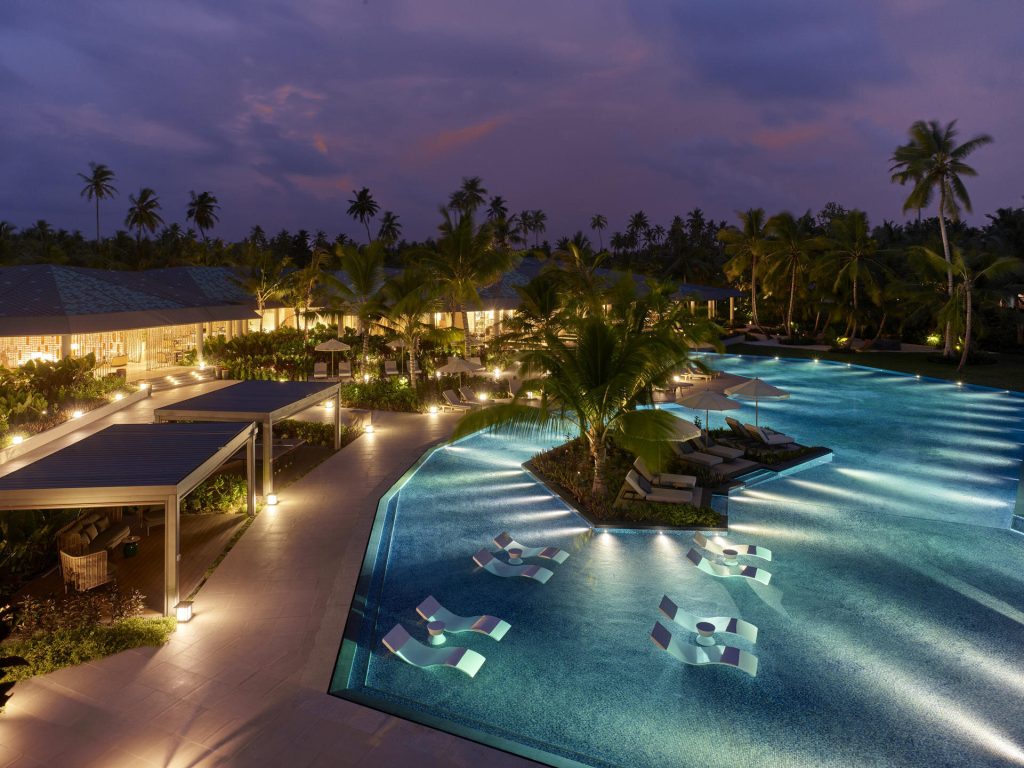 Beleuchteter Pool mit integrierten Liegen, Palmeninsel und Pavillons bei Nacht auf den