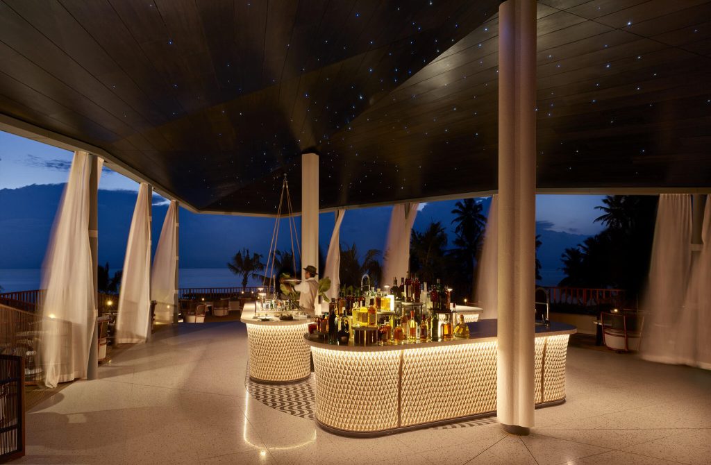 Bar mit gemusterten Theken und Flaschen unter sternförmig beleuchtetem Dach mit Blick auf Palmen und Meer