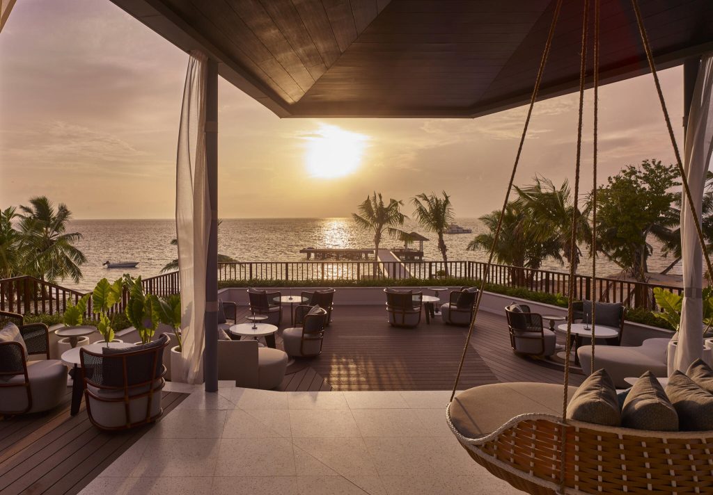 Hotelterrasse mit Holzsesseln, runden Tischen und geflochtener Schaukel bei Sonnenuntergang am Meer