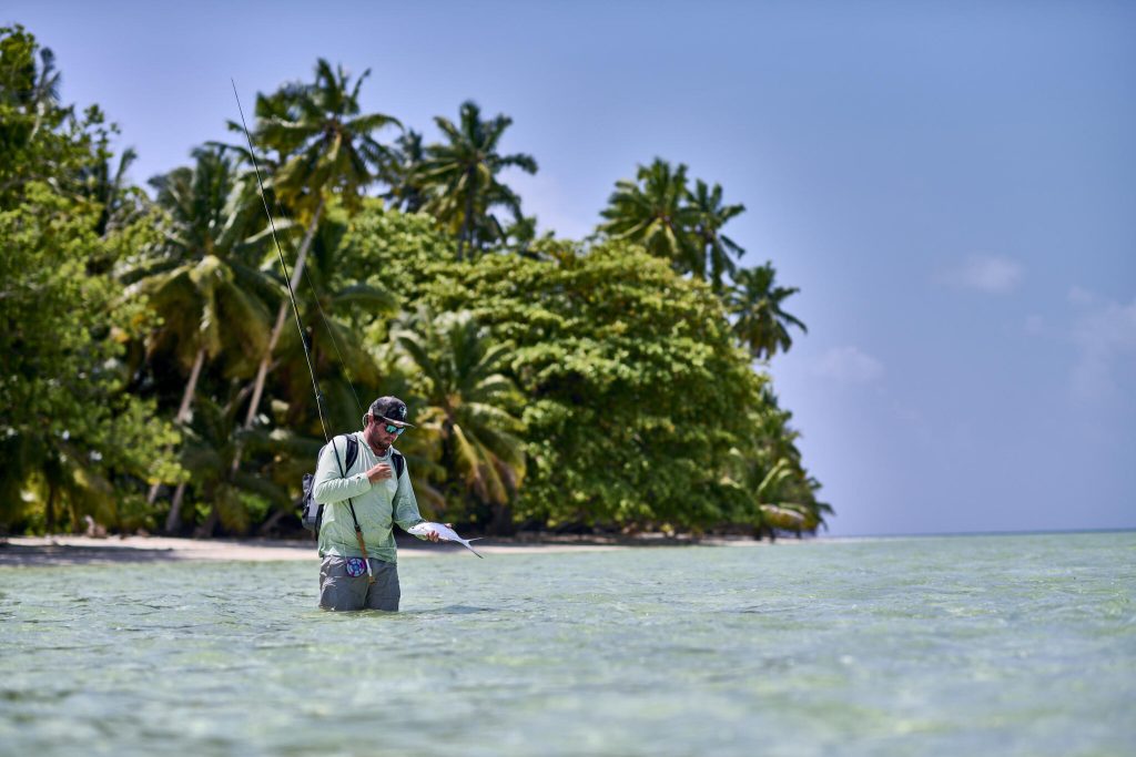 Angler im flachen, klaren Wasser mit Angelrute und Fisch vor palmenbewachsenem Strand auf den