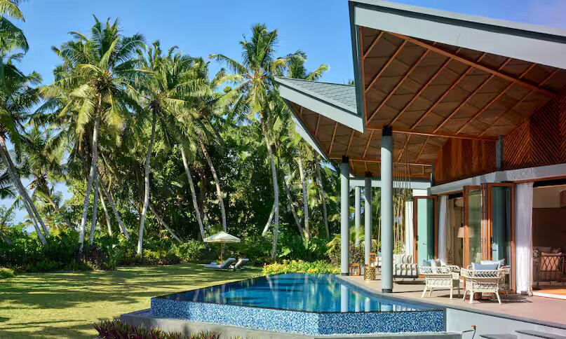 Infinity-Pool mit blauer Mosaikverkleidung vor Holz-Glas-Villa und tropischer Vegetation auf den