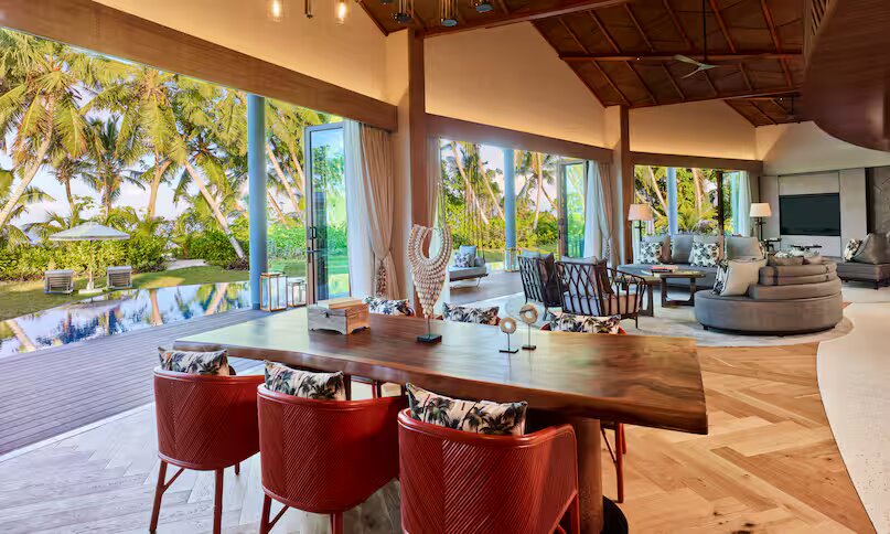 Innenraum der Eagle Ray Pool Villa mit Holztisch, roten Stühlen, Fischgrät-Holzboden und Blick auf Pool und Palmen