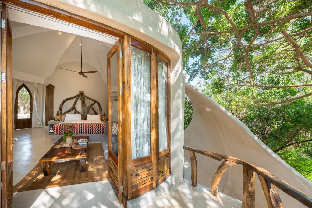 Hell erleuchtetes Zimmer der Villa Alfajiri mit Holzbett, offener Tür zum Balkon und Blick auf grüne Bäume