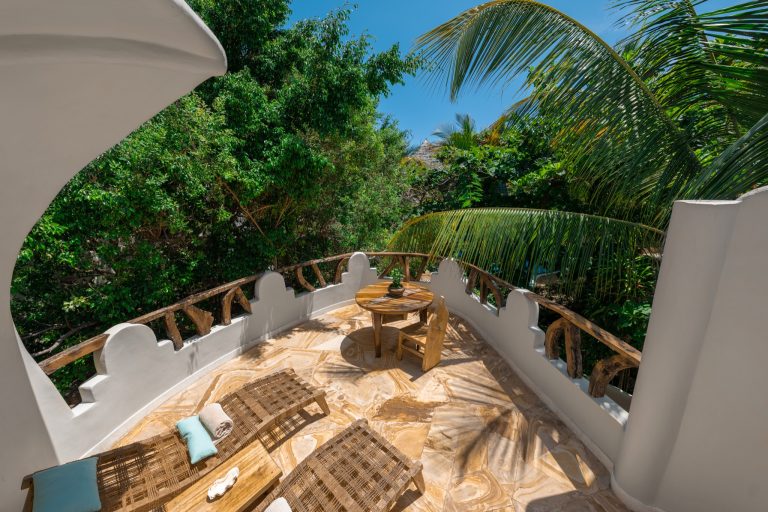 Terrasse der Villa Kimwondo mit zwei Holzliegen, rundem Holztisch und Stühlen sowie grüner Vegetation in Sansibar