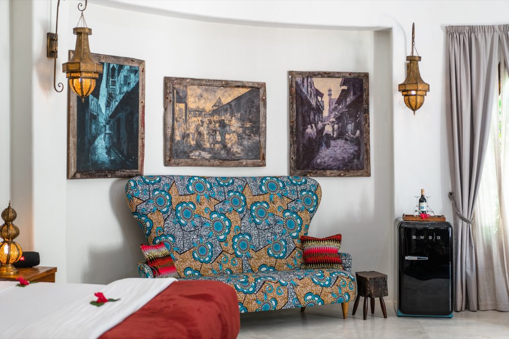 Zimmer der Villa Mawimbi mit buntem Sessel in Blau, Gold und Schwarz, roten Kissen und gerahmten Gemälden an der Wand