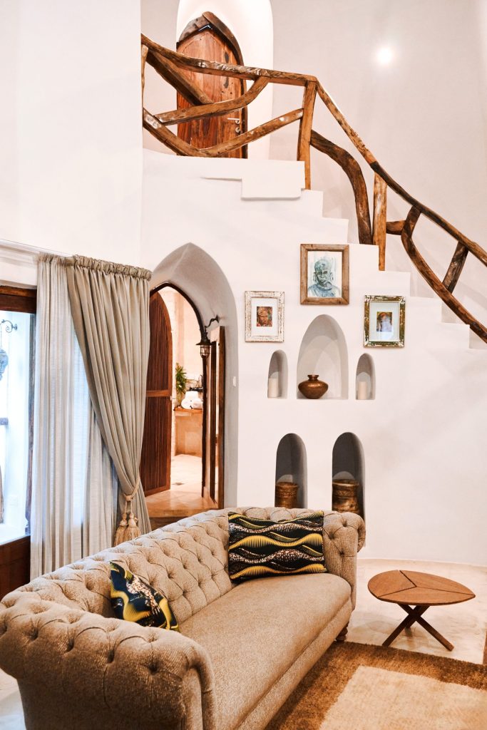 Zimmer der Villa Mbingu in Xanadu Villas mit gepolstertem Sofa, Holztreppe mit Astgeländer und Fenster mit grauem Vorhang