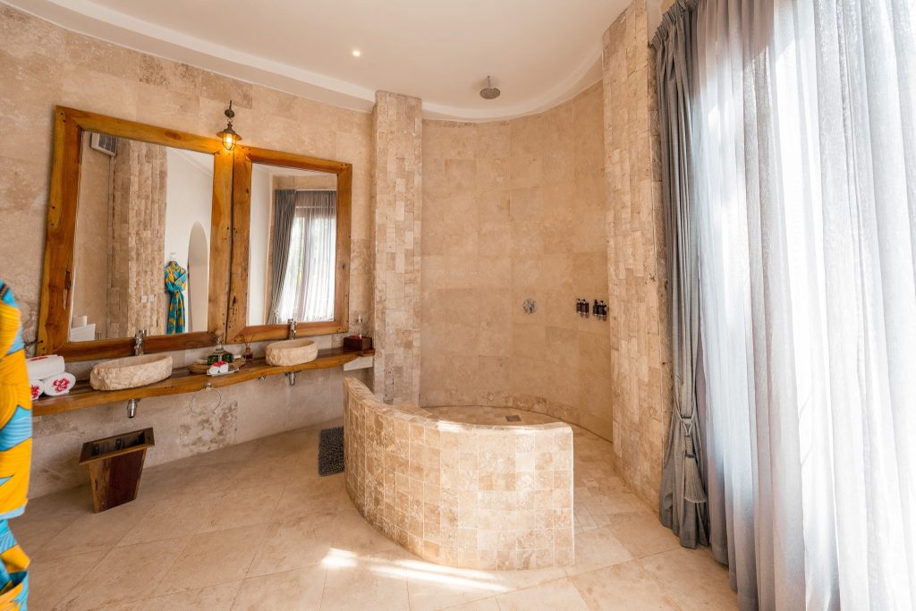 Badezimmer der Villa Mlima in Xanadu Villas mit Natursteinfliesen, zwei Waschbecken und halbkreisförmiger Duschwand