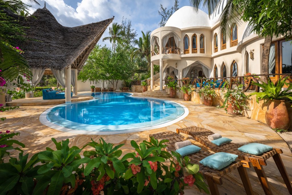 Großer gewundener Pool mit hellbeigen Steinplatten und Strohdächern bei Xanadu Villas Sansibar