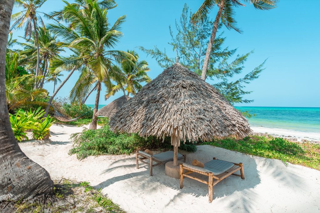 Strandabschnitt mit weißem Sand, Palmen, Pavillon mit Strohdach und zwei Holzliegen mit geflochtenen Auflagen