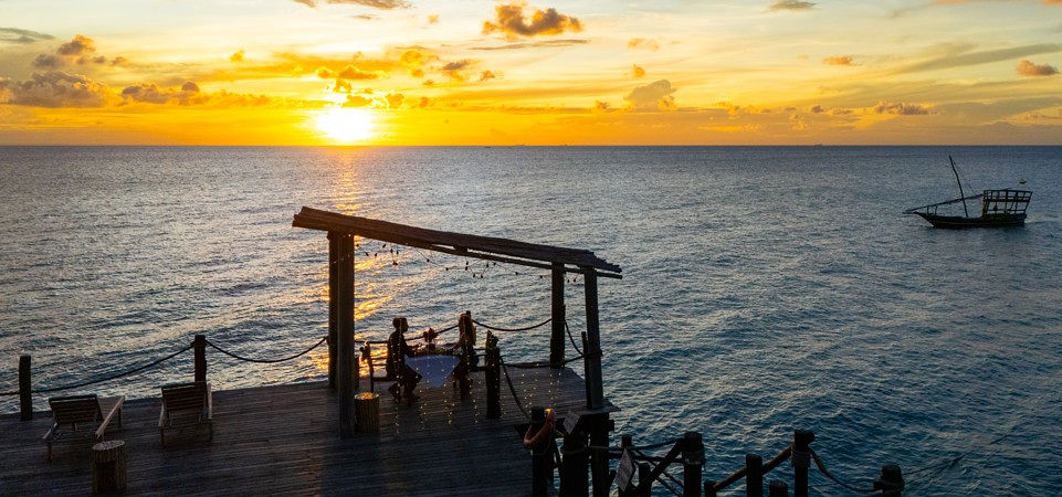 Hölzerner Steg mit Liegestühlen und überdachter Sitzgelegenheit bei Sonnenuntergang am Meer im Zanzi Resort