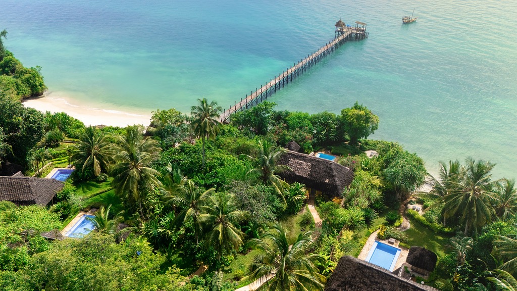 Vogelperspektive auf reetgedeckte Bungalows mit Pools und langem Pier am türkisfarbenen Meer in Sansibar
