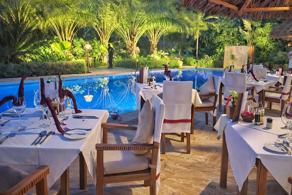 Außenrestaurant mit Holztischen und weißen Tischdecken neben blauem Pool und Palmen bei Dämmerung