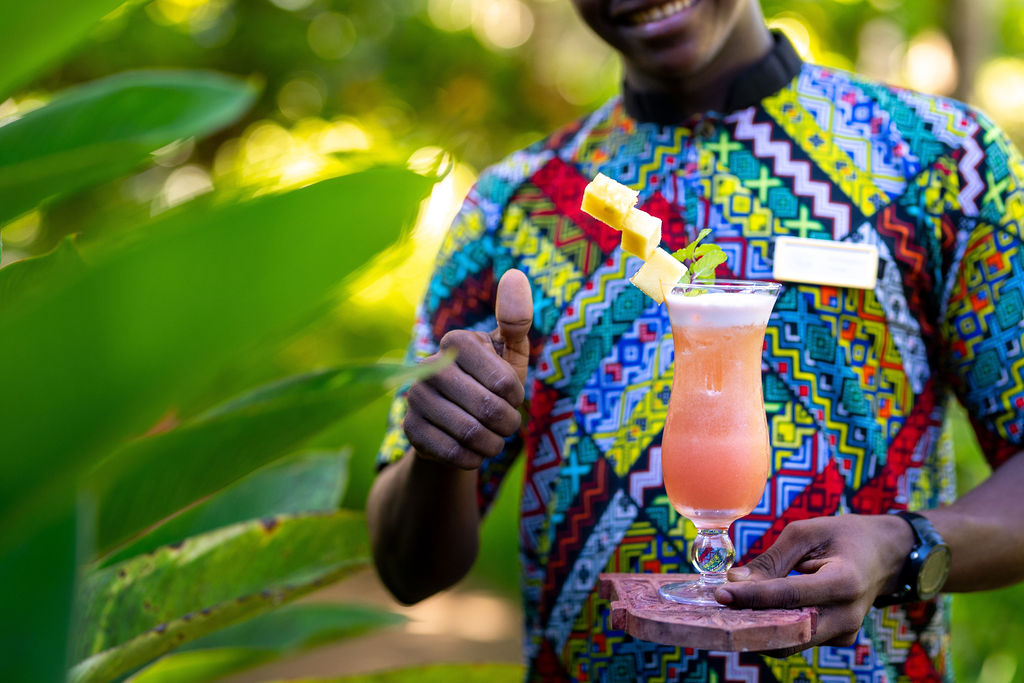 Person im Zanzi Resort Hauptrestaurant hält ein Glas mit cocktailartigem Getränk und Fruchtgarnitur vor grüner tropischer.