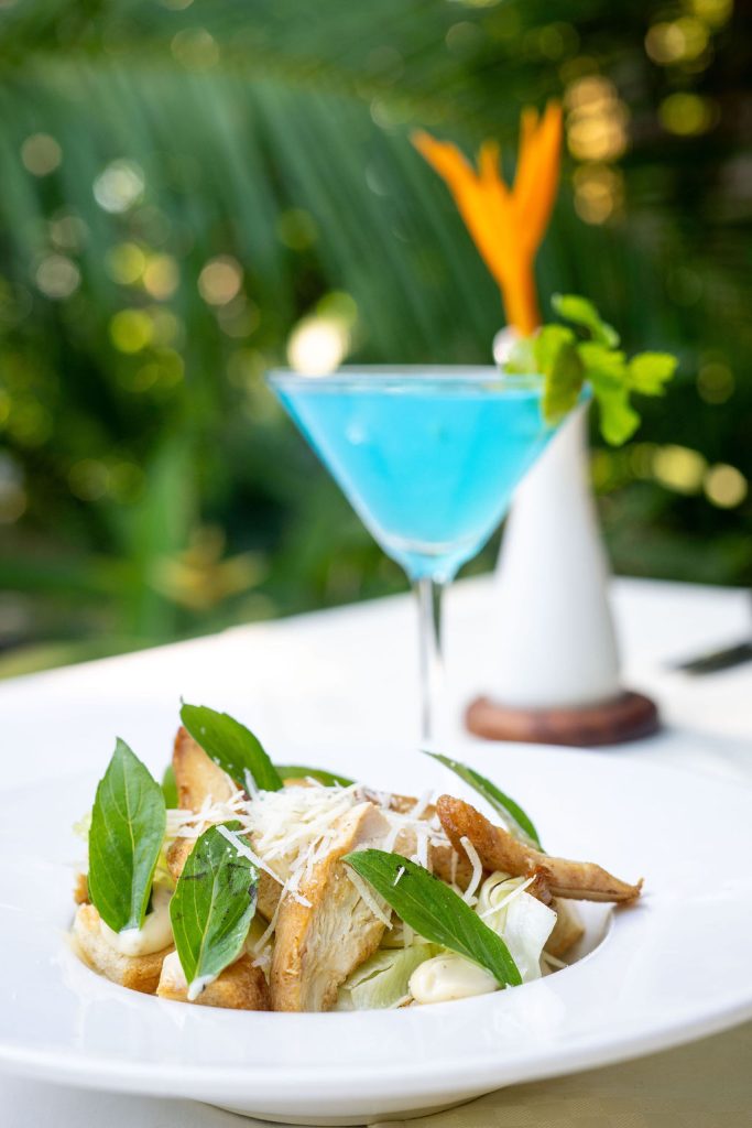 Teller mit grünem Salat und geriebenem Käse auf weißem Tischtuch, blauer Cocktail mit mintgrüner Garnitur vor grüner.