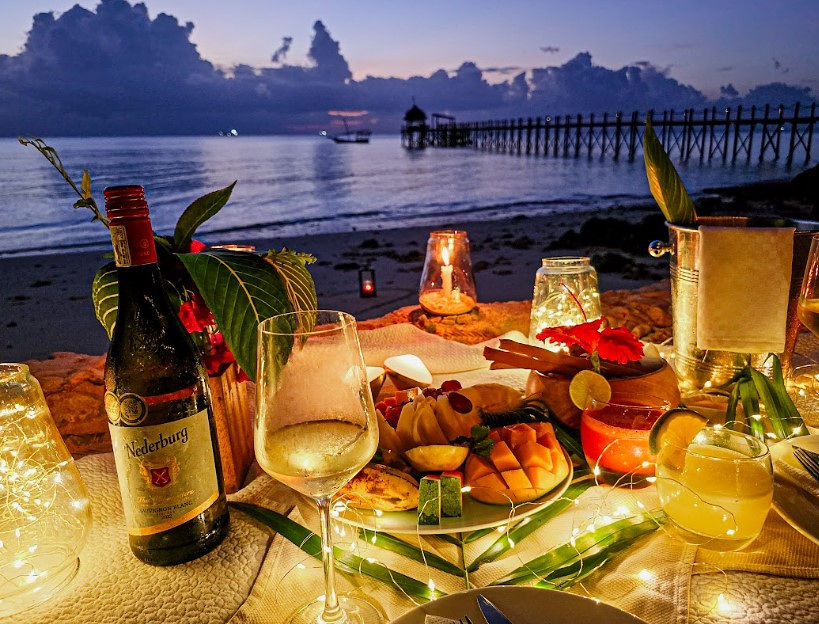 Gedeckter Holztisch mit Wein, Gläsern und Früchten am Strand bei Dämmerung im Restaurant Hauptrestaurant