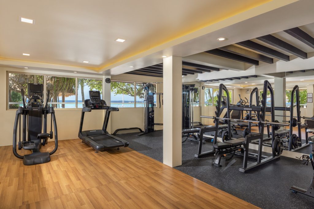 Moderner Fitnessraum mit Crosstrainer, Laufband, Kraftstation, Holzlaminatboden und großen Fenstern zum Meer
