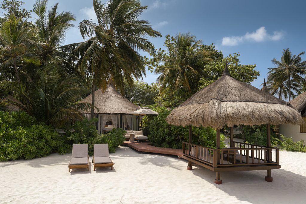 Strandbereich der Banyan Tree Vabbinfaru Beachfront Pool Villa mit Holzliegen, Strohdach und Palmen im Hintergrund