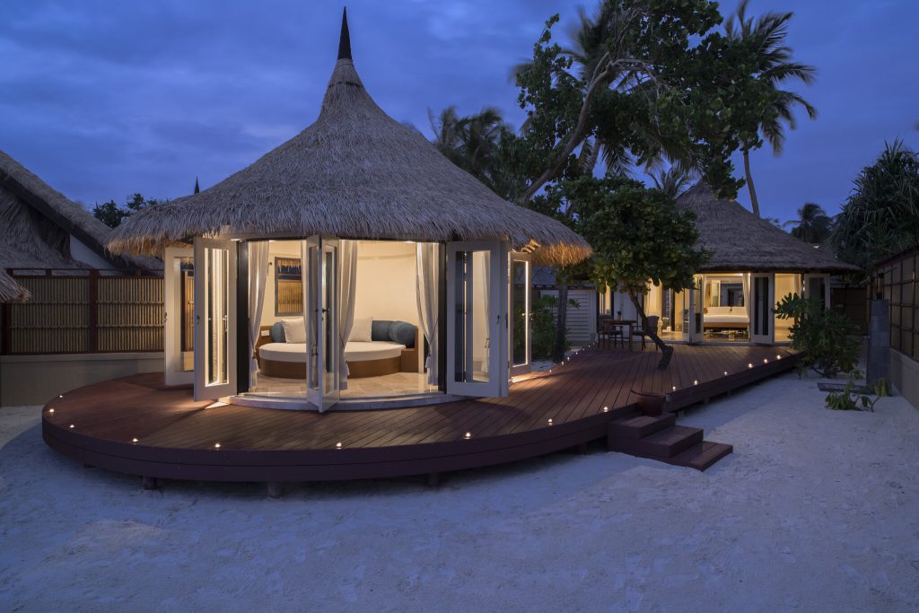 Grand Beachfront Pool Villa mit Strohdach, Holzdeck und geöffneten Türen bei Dämmerung auf den