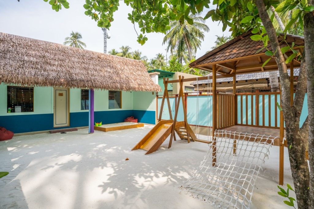 Außenbereich im Dhawa Ihuru Kids Club mit Holzspielplatz, sandbedecktem Boden und türkis-blauem Gebäude