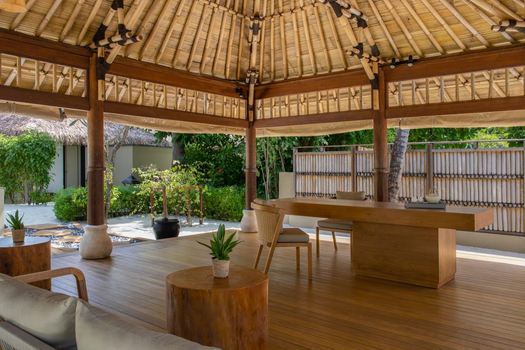 Offene Sitzecke im Dhawa Ihuru Wellness Spa mit Holztisch, Bambusdach und Blick auf grünen Garten