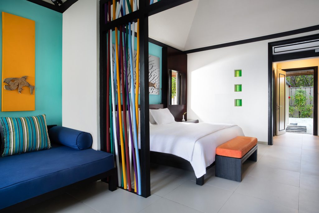Modernes Hotelzimmer mit blauem Sofa, weißem Bett, orangefarbenem Hocker und Blick auf grüne Außenterrasse im Dhawa Ihuru