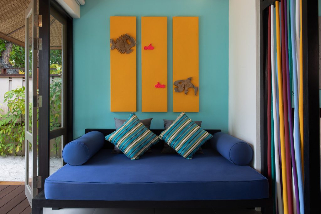 Innenbereich der Beachfront Jet Pool Villa mit blauem Sofa, gestreiften Kissen, gelben Holzpaneelen und Blick auf grüne.