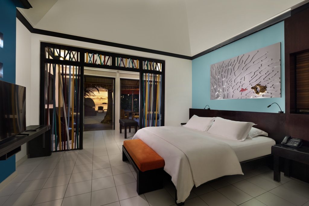 Hotelzimmer der Beachfront Sunset Villa mit Doppelbett, blau-weißer Wand, Holzmöbeln und Sonnenuntergang auf den