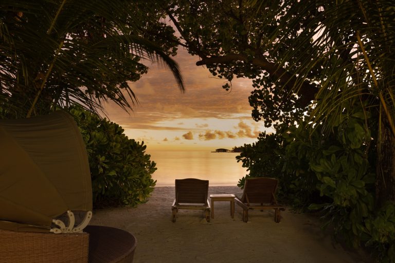 Ruhiger Strandabschnitt mit zwei hölzernen Liegestühlen, Tisch und Pflanzen bei Sonnenuntergang
