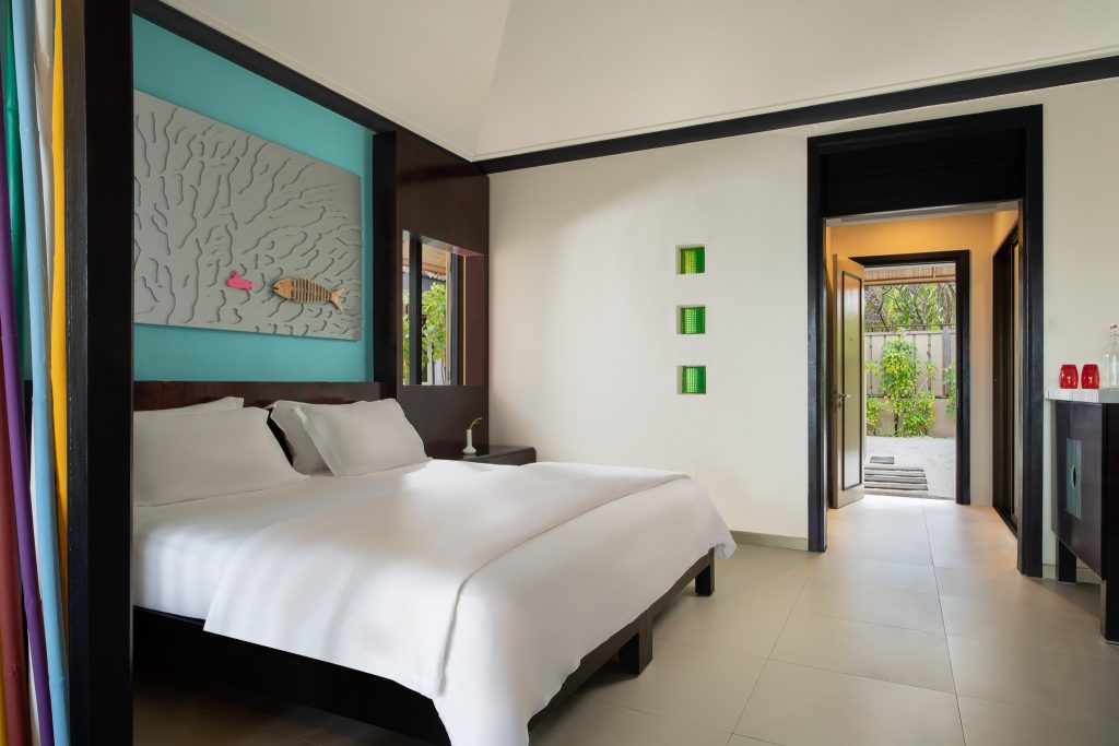 Modernes Hotelzimmer Dhawa Ihuru Beachfront Villa mit großem Bett, Holzrahmen und türkisfarbenem Kunstwerk über dem Bett