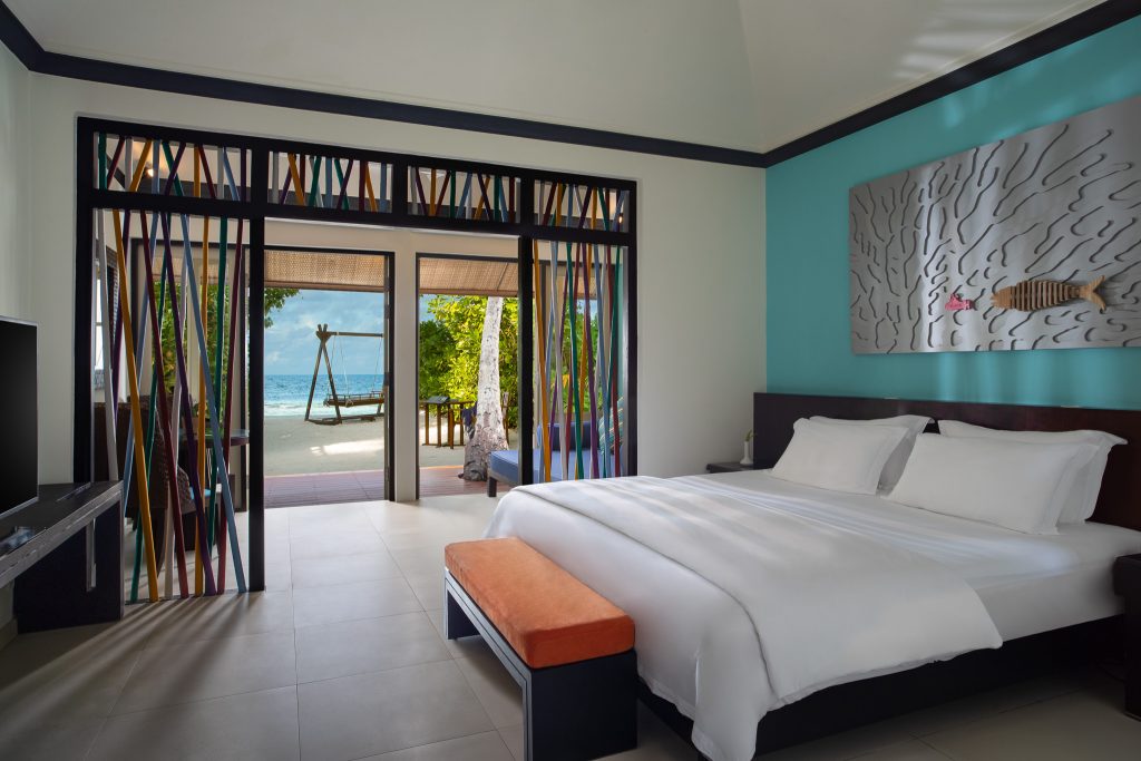Modernes Zimmer der Beachfront Villa im Dhawa Ihuru mit großem Bett, türkisfarbener Wand und Blick auf Terrasse und Meer