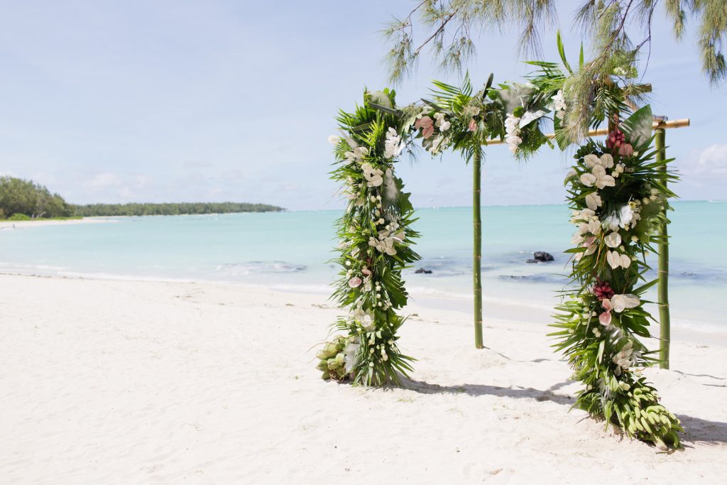Strand mit Bambus-Torbogen, geschmückt mit grünen Blättern, weißen und rosa Blumen, feinem Sand und blauem Meer.