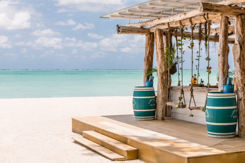 Offene Strandbar aus Holz mit zwei türkisfarbenen Fässern auf Holzpodest am weißen Sandstrand und Meer im Hintergrund