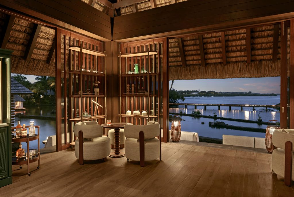 Hotelzimmer mit Holzmöbeln, zwei beige Sessel, runder Holztisch, Wandregal mit Vasen und Blick auf Wasser