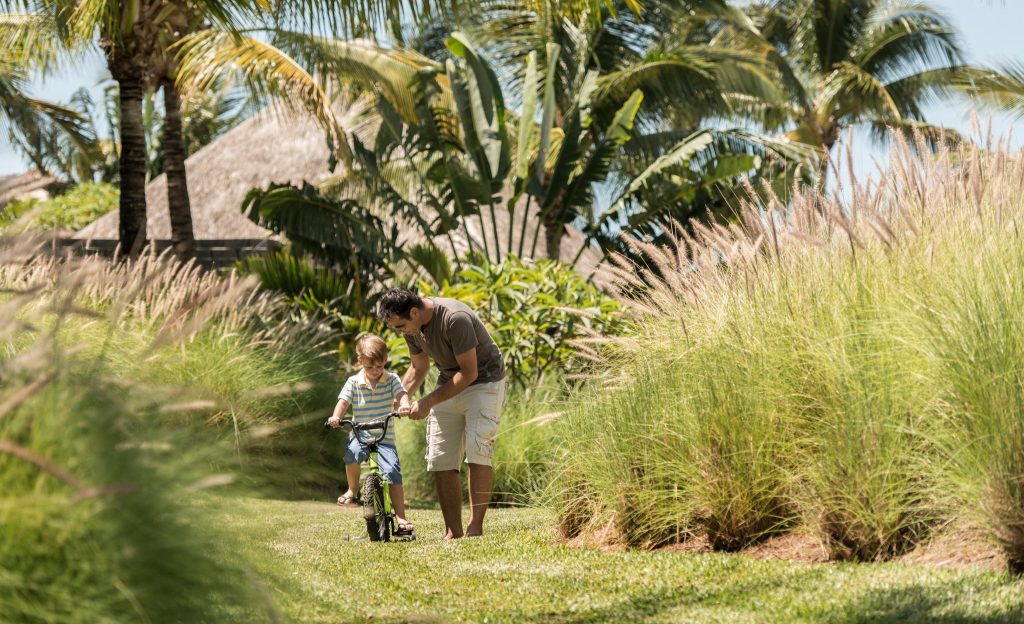 Erwachsener und Kind auf schmalem Rasenpfad im Garten des Four Seasons Resort Mauritius At Anahita mit Fahrrad