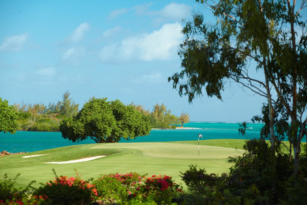 Golfplatz mit grünem Fairway, Sandbunker und bunten Blumen vor türkisblauem Meer und Bäumen am Ufer
