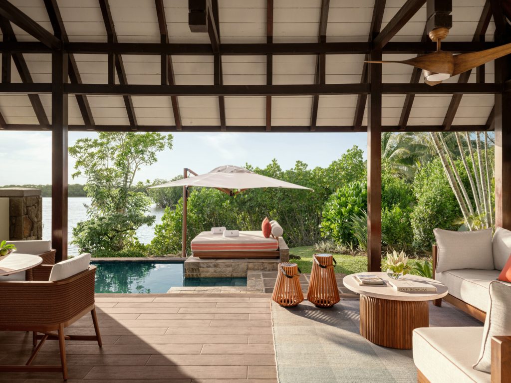 Offene Terrasse eines Hotelzimmers mit Holzmöbeln, Pool, Sonnenbett und grüner Vegetation bei Tageslicht