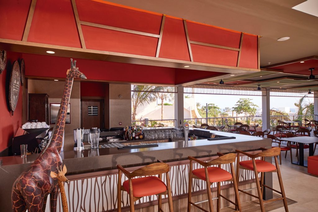 Barbereich im Hotel Riu Jambo mit Holzbarhockern, großer Giraffenfigur und Blick auf Außenterrasse und Palmen