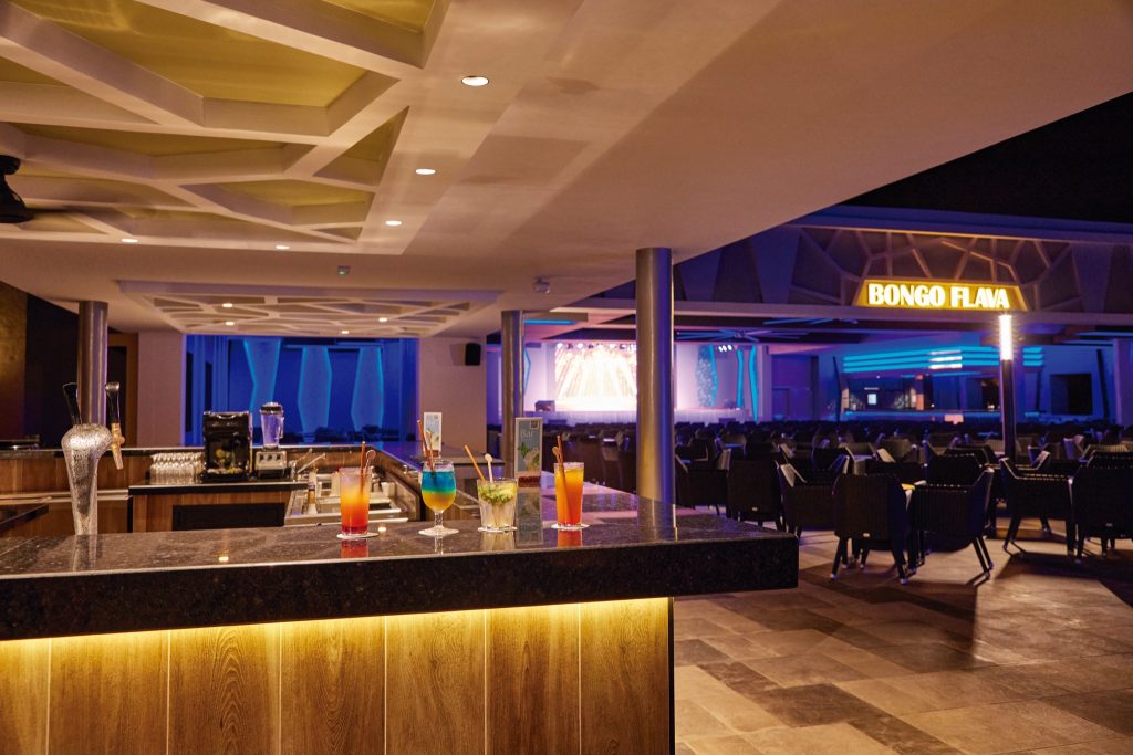 Moderne Bar im Hotel Riu Jambo mit dunkler Stein-Theke, Holzdetails, bunten Cocktails und Bühne im Hintergrund