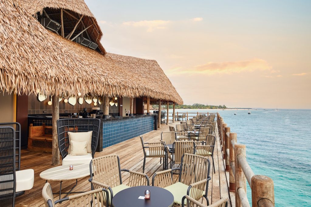 Terrasse der Bar Hotel Riu Jambo mit Holzdeck, Metall-Holz-Tischen und Strohdach über dem Meer bei Sansibar