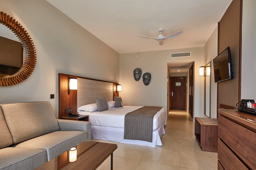Modernes Hotelzimmer im Hotel Riu Jambo mit großem Bett, grauem Sofa, Holztisch und dekorativen Masken an der Wand