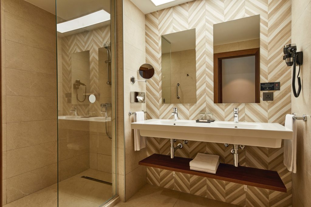 Modernes Badezimmer im Hotel Riu Jambo mit beigen Fliesen, Doppelwaschbecken, großer Spiegel und gläserner Duschwand