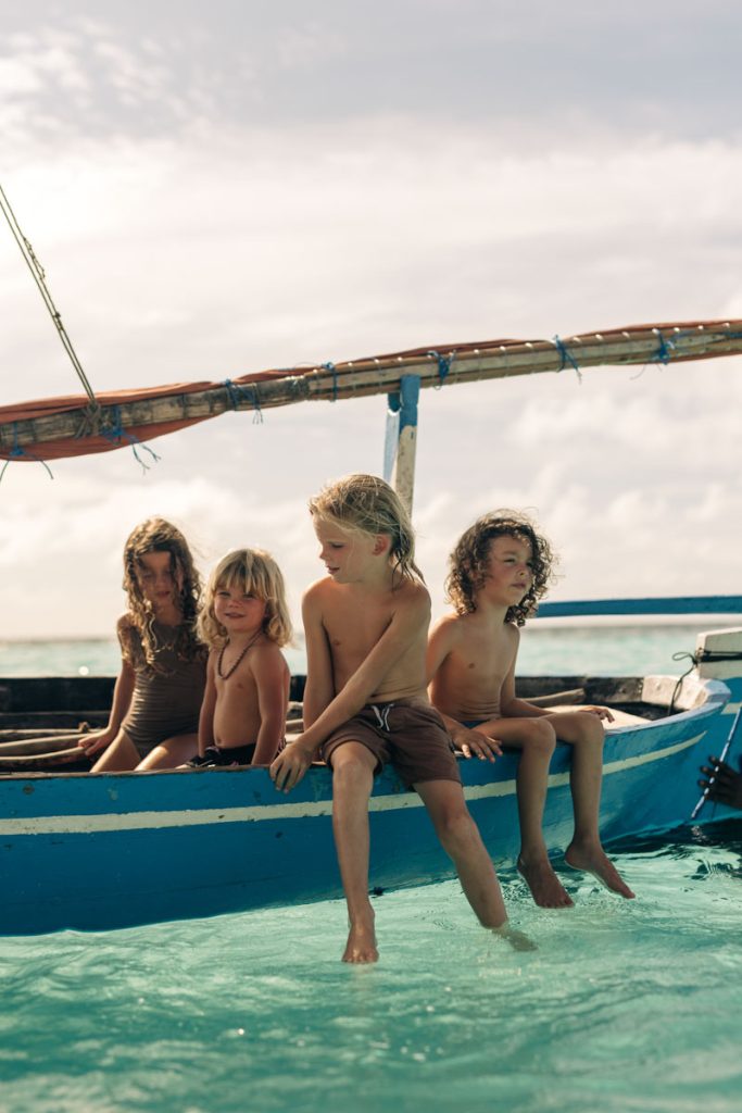 Vier Kinder sitzen auf einem blau-weißen Holzboot auf türkisfarbenem Meer vor leicht bewölktem Himmel