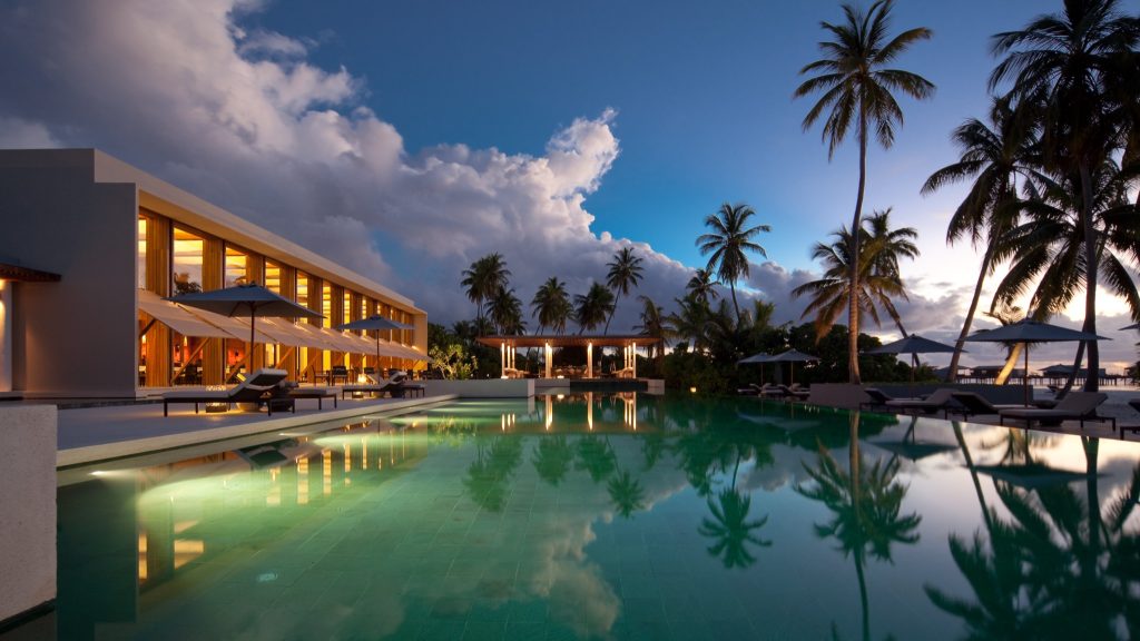Großer Pool vor modernem Hotel mit Glas, Beton und Holzrahmen, Palmen und Himmel spiegeln sich